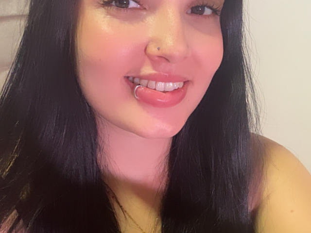 SarahGood with black hair, nose ring, and lip ring, smiling with pinkish cheeks and shiny lips, showing her teeth. Her ass is not visible.
