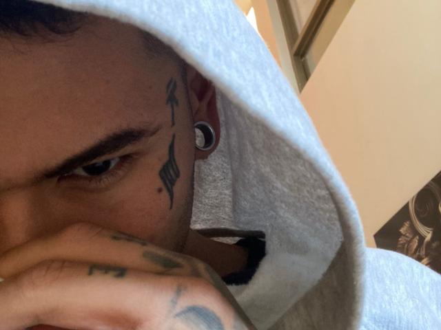 JamesInked trägt eine graue Kapuzenjacke, die seine Nase bedeckt, hat Tattoos, schwarze Ohrpiercings und einen ernsten Gesichtsausdruck. Man sieht sein blaues Auge und seine tätowierte Hand..