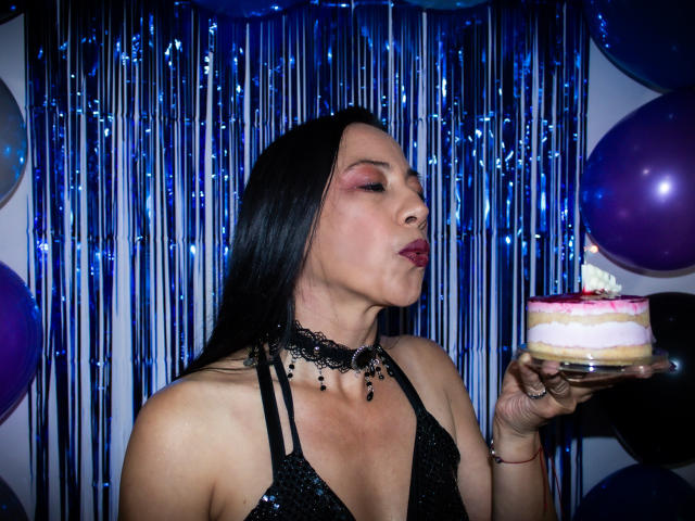 NoemiPrettiest with long black hair, wearing a black sequined halter top and black lace choker, blows pink lipstick kiss at a pink and white cake. Blue metallic streamers.