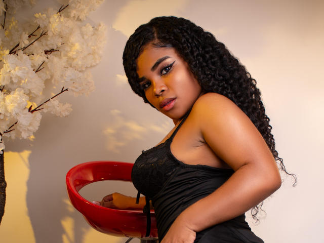 NahiaraReina with curly black hair, wearing a black lace dress, holding a red bowl, showing her ass, standing next to white cherry blossoms, looking at the camera.