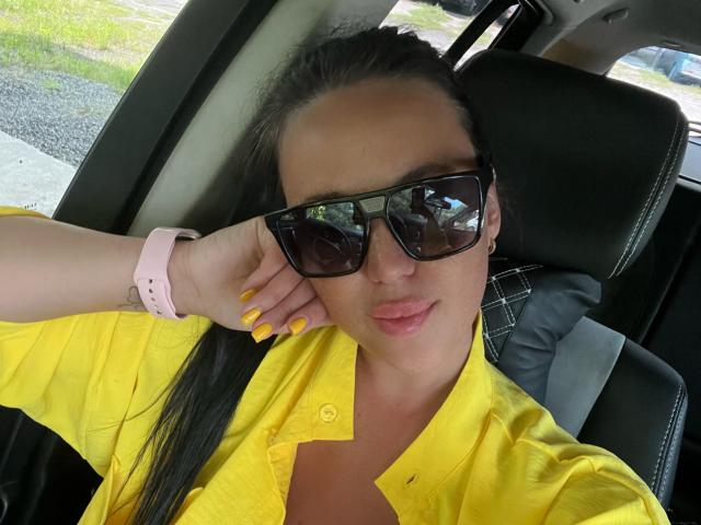 FinessiaLovely, con una camicia gialla, occhiali da sole neri, un orologio rosa, unghie gialle e capelli neri, seduta in macchina, che ostenta il suo seno..