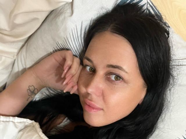 FinessiaLovely lying in bed, dark hair, green eyes, light skin, small tattoo on her wrist, slightly smiling, hand under chin, white pillow and blanket.