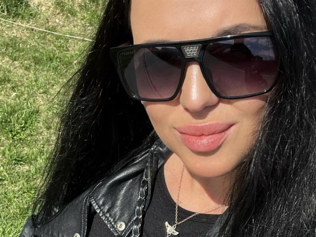 FinessiaLovely, con i capelli neri, indossa occhiali da sole neri, una giacca di pelle nera e una camicia nera, ha labbra rosa lucide, una collana d'argento con una piccola croce ed è sorridente..