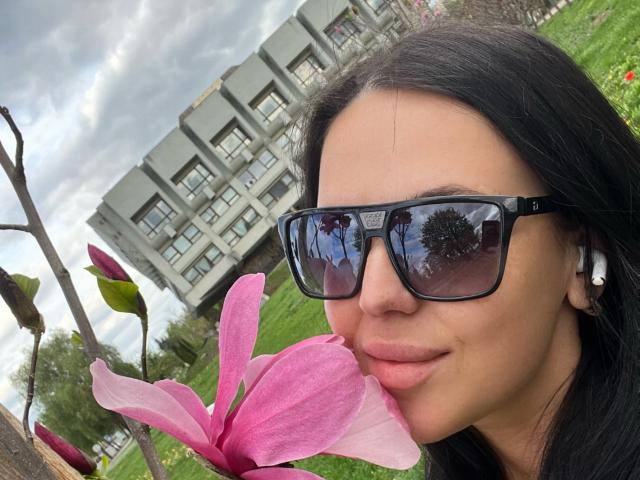 FinessiaLovely, con i capelli neri, che indossa occhiali da sole, tiene vicino alla bocca un fiore di magnolia rosa, in piedi di fronte a un edificio residenziale grigio, cielo nuvoloso, auricolari bianchi..