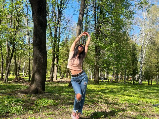 FinessiaLovely, con lunghi capelli castani, piccoli seni, indossa una maglietta rosa e jeans strappati, braccia alzate, in piedi in una foresta, con la luce del sole che filtra tra gli alberi..