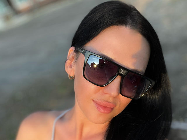 FinessiaLovely with long black hair, wearing dark sunglasses, and a small hoop earring, has a subtle smile and a light tan, showing her full lips and a cute nose.