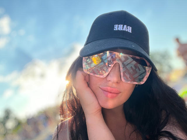 FinessiaLovely with long black hair, wearing a black hat and pink reflective sunglasses, pouting her pink lips, sunlight shining on her face, hand resting on cheek.