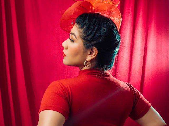 Khloe Lawson, avec des cheveux foncés, porte un pull rouge à col roulé, un chapeau rouge-orange orné de plumes, des boucles d'oreilles dorées et regarde sur le côté. L'arrière-plan est rouge. Les seins, les mamelons et les fesses ne sont pas visibles..