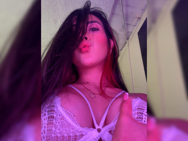 AnyaJay with pouty pink lips, dark hair, white lace bra, showing her tits, pink and purple lighting, holding a mirror, long nails, necklace, and a slight red tint in hair.