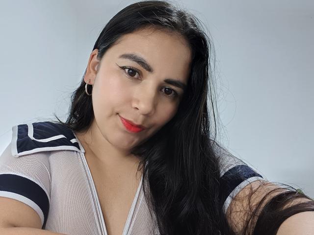 SofiaCopers has long black hair, red lipstick, and wears a sheer white top with navy and white stripes. She's smiling slightly, looking at the camera.