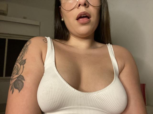 AlexaLip with her tits in a white ribbed tank top, open-mouthed, glasses on, tattoo on her left arm, brown hair, pearl earrings, in a dimly lit room.