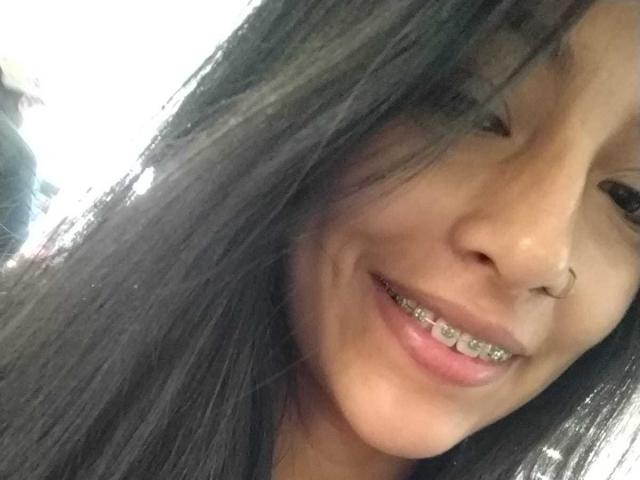 NoremCute's face, showing a smile with a nose ring and metal braces on her teeth, has long black hair covering part of her face. She's looking slightly to the side.