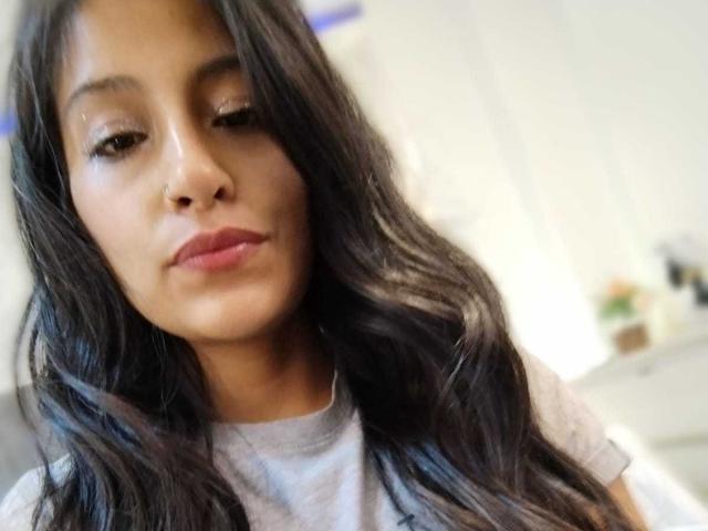 NoremCute has long, wavy black hair, brown eyes, and pink lips. She's wearing a white shirt, with a subtle nose ring on her left nostril. She's looking down.