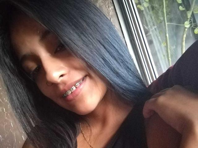 NoremCute has long black hair, a nose ring, and metal braces on her teeth, smiling softly with her hand near her neck, standing by a window with green vines.