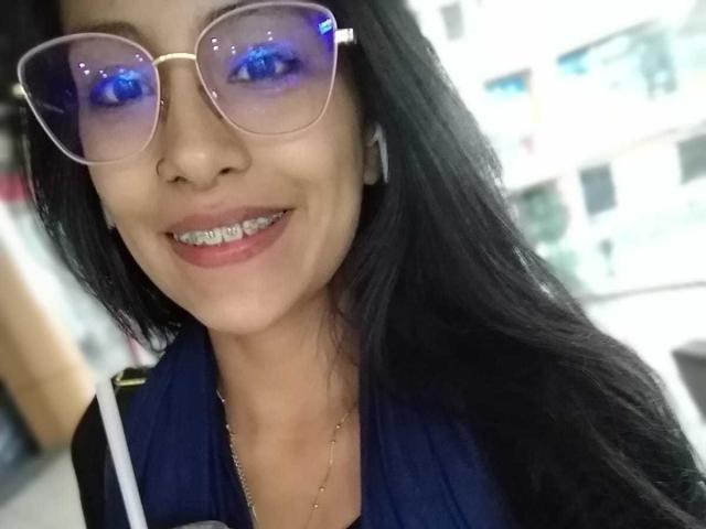 NoremCute has blue-tinted glasses, black hair, and a smile showing metal teeth. She's wearing a blue shirt with white straps, and has a gold necklace.