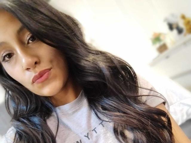 NoremCute has long, wavy black hair, light brown skin, and full pink lips, wearing a white t-shirt with small black crosses. She's looking slightly to the right.