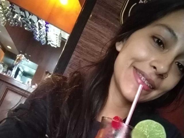 NoremCute with long dark hair, nose ring, and a slight smile, sipping through a straw from a drink with a lime and cherry, wearing a black top.