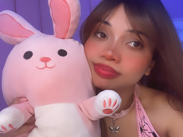 LiliMays with pink bunny stuffed animal, holding it against her face. She's got long brown hair, red lipstick, pink collar, and a pink halter top. Her tits are small.