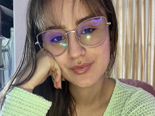 NinaBryons with glasses reflecting blue and purple lights, nose ring, and green sweater, smirking with hand on cheek, brown hair, and shiny lips. Her ass is not visible.