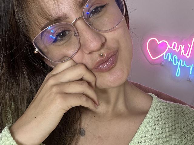 NinaBryons with glasses, nose piercing, and lip ring, pouting, wearing a green knit sweater, hand under chin, small tattoo on chest, neon heart sign in background.