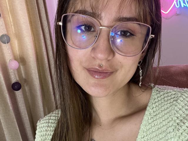 NinaBryons with glossy lips, nose piercing, and big round glasses, wearing a green knitted top, smiling at the camera, brown hair, and pink neon light in the background.