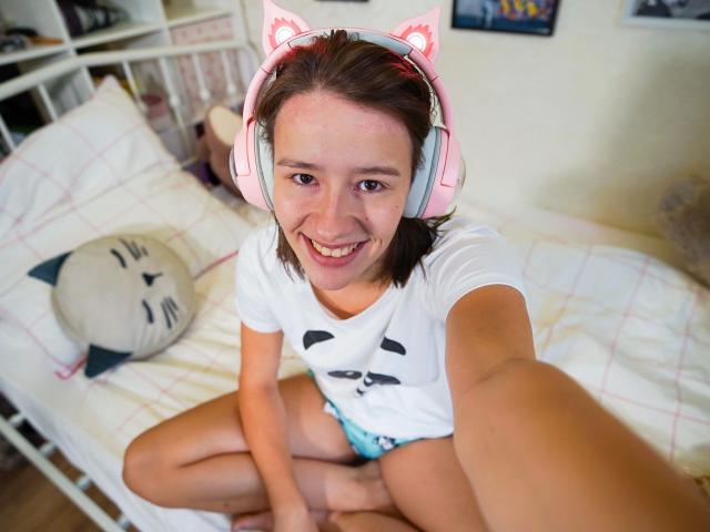 LenaBlush: bellissima, con orecchie da gatto rosa, sorridente, maglietta bianca, slip blu, seduta sul letto, che si fa un selfie, letto bianco con cuscino a forma di gatto, capelli castani, pelle chiara, carina, allegra..