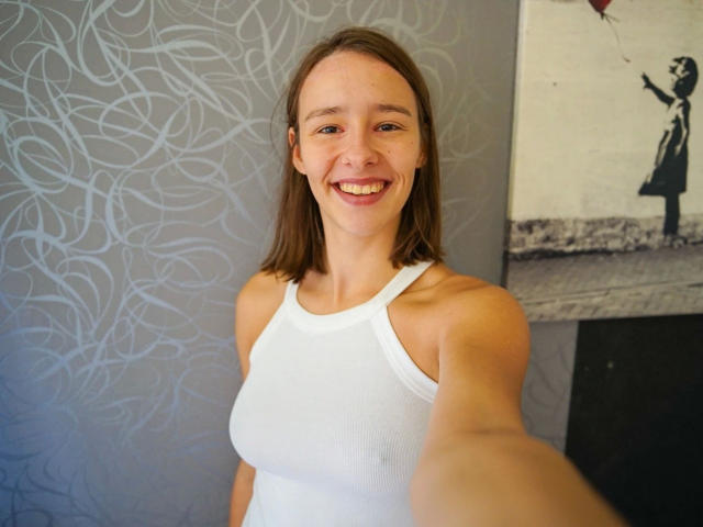 LenaBlush – attraktiv mit mittlerer Brustgröße, lächelnd, trägt ein weißes Tanktop, macht ein Selfie, braunes Haar, blaue Augen, helle Haut, steht in einem Raum mit abstrakt gemustertem Tapeten..