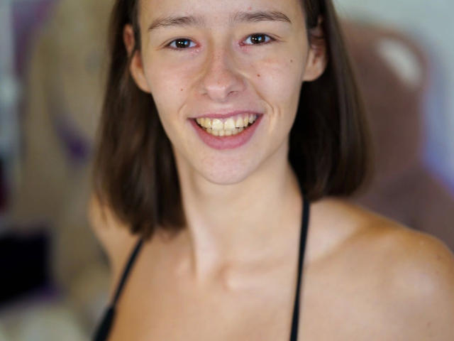 LenaBlush: una ragazza attraente con capelli castano di lunghezza media, che sorride mostrando i denti, indossa un top nero con spalline, ha una carnagione chiara e piccole lentiggini sul naso..