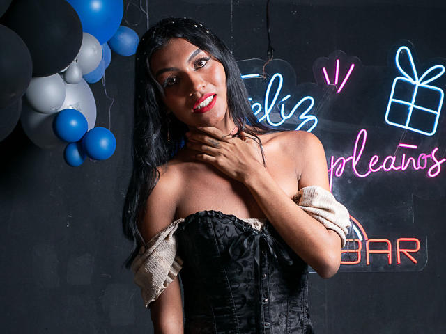 KattaleyaHorny with dark skin, long black hair, red lipstick, black off-shoulder corset, and beige sleeves, touching her neck, smiling, neon signs in the background.