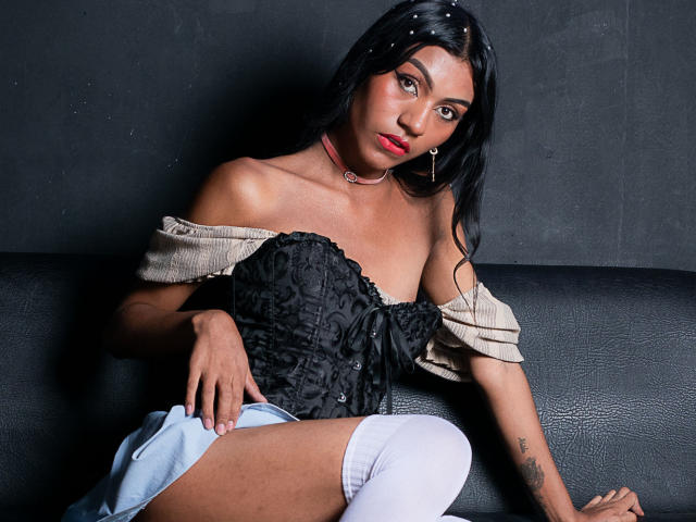 KattaleyaHorny with black lace corset, off-shoulder beige top, white thigh-high socks, and blue shorts, sitting on black leather couch, looking at camera with red lipstick.