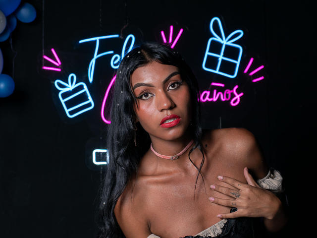 KattaleyaHorny with dark skin, long black hair, red lipstick, choker, off-shoulder top, hand on chest, neon "gifts ans" in background, blue and pink neon lights.