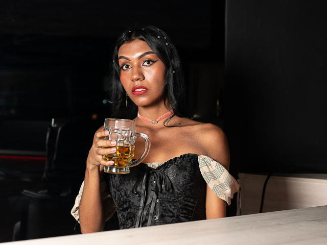 KattaleyaHorny with dark skin, black lace off-shoulder top, holding a beer mug, red lipstick, black hair, serious expression, in a dimly lit bar, showcasing her tits.