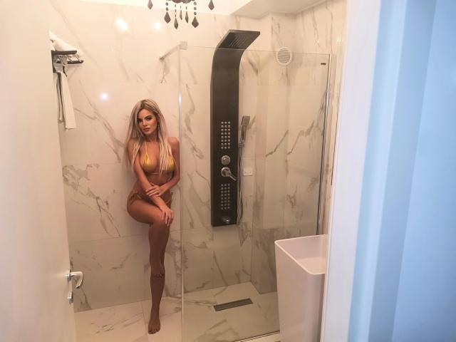 AshleyWise staat in een marmeren douche, gekleed in een geel bikinietje dat haar grote borsten en kleine billen accentueert. Ze heeft lang blond haar en een sexy, zelfverzekerde uitstraling..