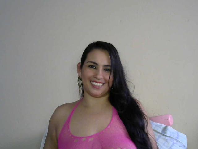 ZoeNava, com cabelos longos e pretos, usando um top de malha rosa com alças, sorrindo, mostrando seios grandes, usando brincos de argola, sentada em frente a uma parede bege..