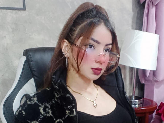 AshleyMiler with brown ponytail, pink glasses, red lipstick, black top, black patterned jacket, gold necklace, and earrings, sitting in black chair, white lamp on right.