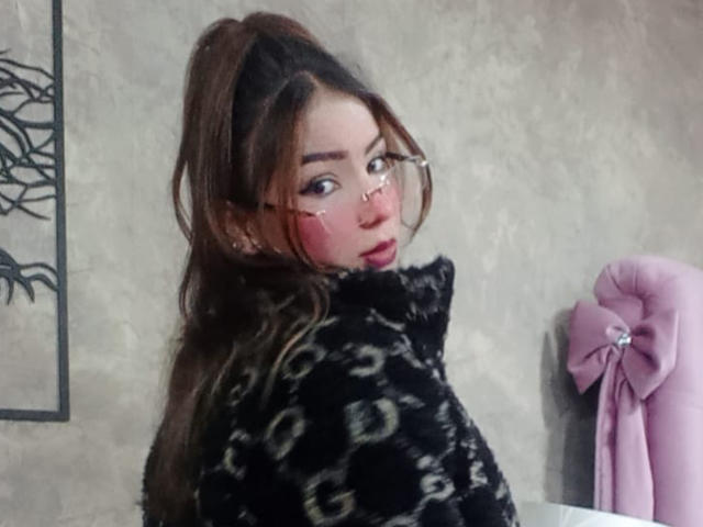 AshleyMiler with long brown hair in a ponytail, wearing glasses and a black patterned fur coat, has pink blush on her cheeks, looking over her shoulder. Pink bow on wall.