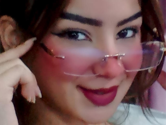 AshleyMiler with a red nose clip, wearing glasses, and pink blush on her cheeks, smiling with dark red lipstick, and her fingers near her face.
