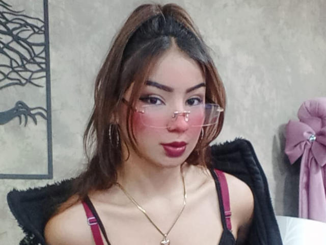 AshleyMiler with pink glasses, red lipstick, and hoop earrings, wearing a black and red top, has her tits slightly visible, and a gold necklace with a heart pendant.