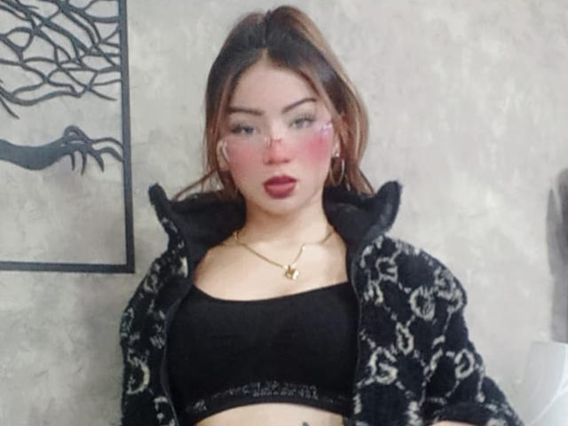 AshleyMiler with brown hair, wearing a black sports bra and floral patterned jacket, has pink eyeshadow, red lipstick, and a gold necklace, showing her tits.