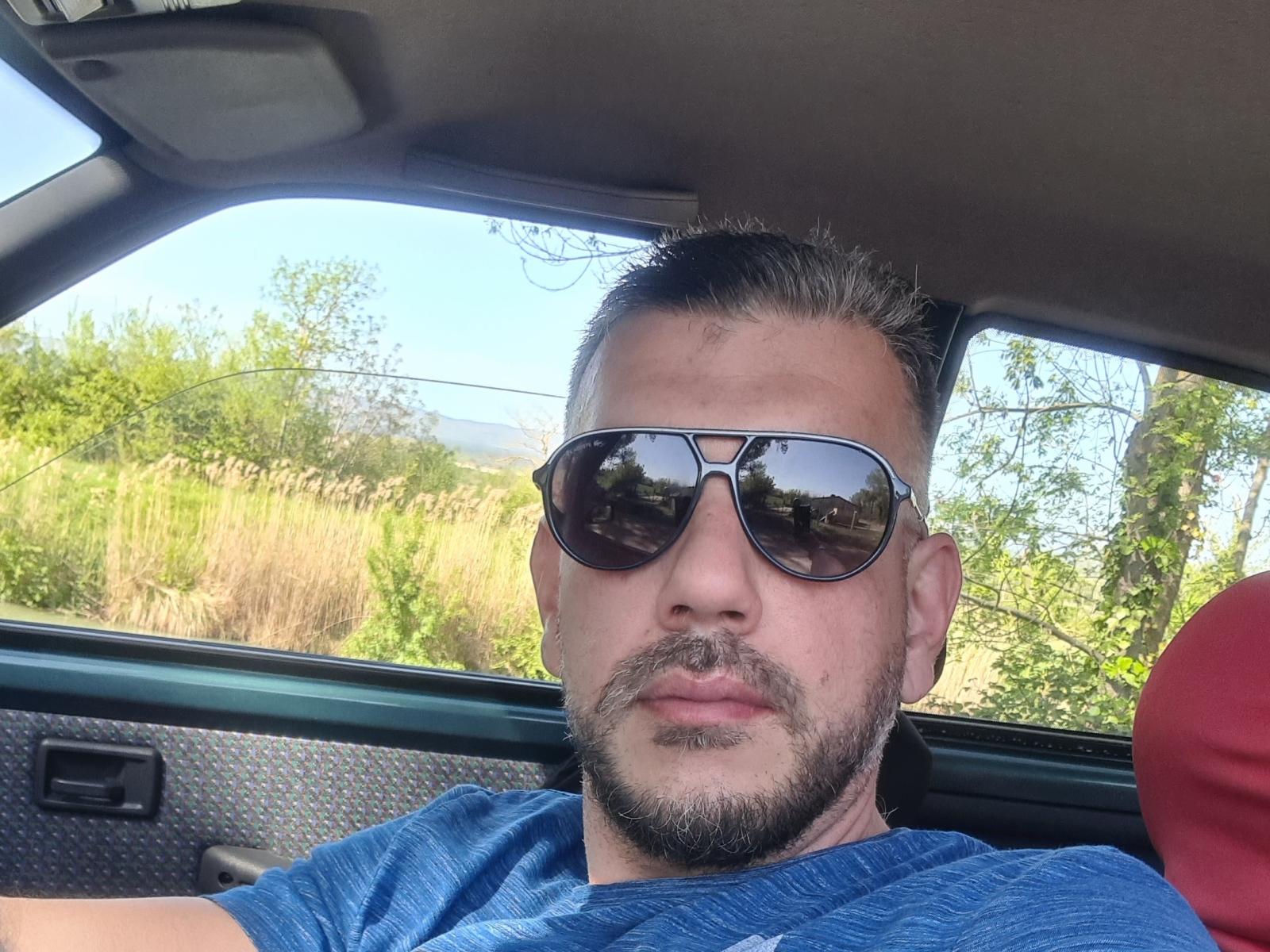 SexyMarcoHott with a beard, wearing dark sunglasses and a blue shirt, looking serious while driving a car. His reflection is in the sunglasses.
