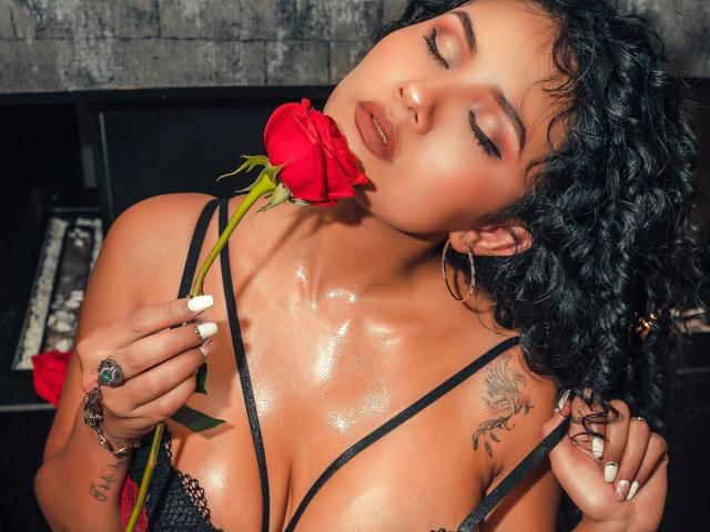 CanelaLebrad with shiny, oiled-up tits, holding a red rose to her nose, eyes closed, curly black hair, hoop earrings, tattoo on her right shoulder, wearing black lingerie.
