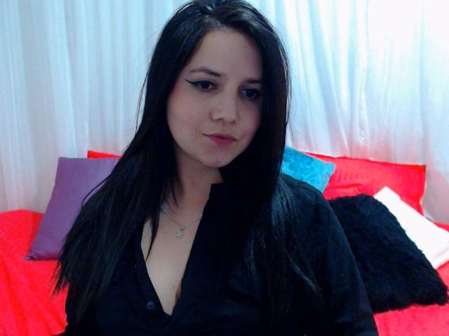 Kkatalina with black hair, wearing a black shirt, showing her small tits, sitting on a red bed with pillows, looking at the camera with a slight smile.