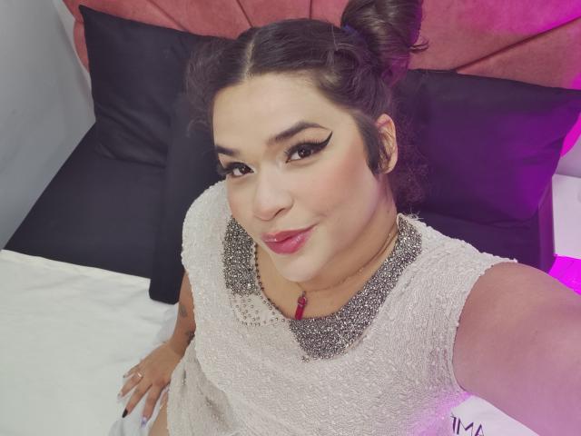 AntonelaPark with puffy brown hair in buns, wearing a sparkly beige dress, bold eyeliner, pink lipstick, and a necklace, sitting on a bed with black and pink pillows.