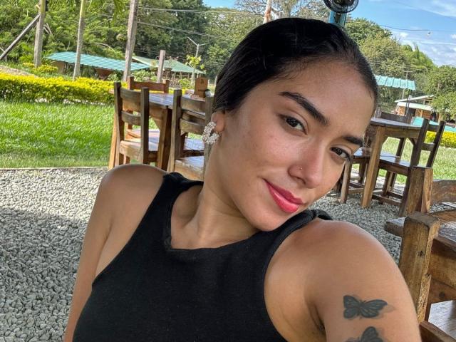 SharonKlart with dark hair in a black tank top, pink lipstick, small nose ring, butterfly tattoo on right arm, smiling, outside on gravel, wooden chairs in background.