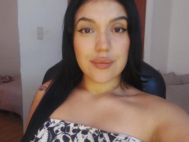 LauraRichis with black hair, brown eyes, and light skin, wearing a strapless top with a black and white skull pattern, sitting in a chair, smiling slightly.