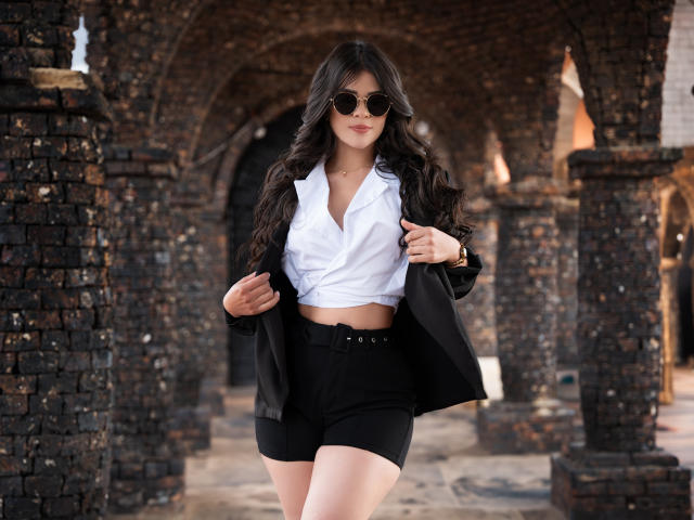 PaulinaValley with long dark hair, wearing round sunglasses, white button-up shirt, black jacket, and black shorts, holding jacket open, standing in brick archway. Her ass is visible.
