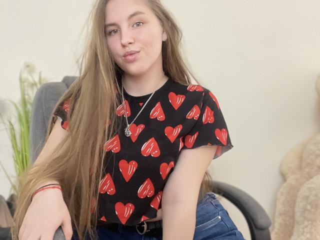 Dooris with long brown hair, pouty lips, wearing a black heart-print shirt and blue jeans, sitting in a chair, necklace, and red bracelet, looking at the camera.