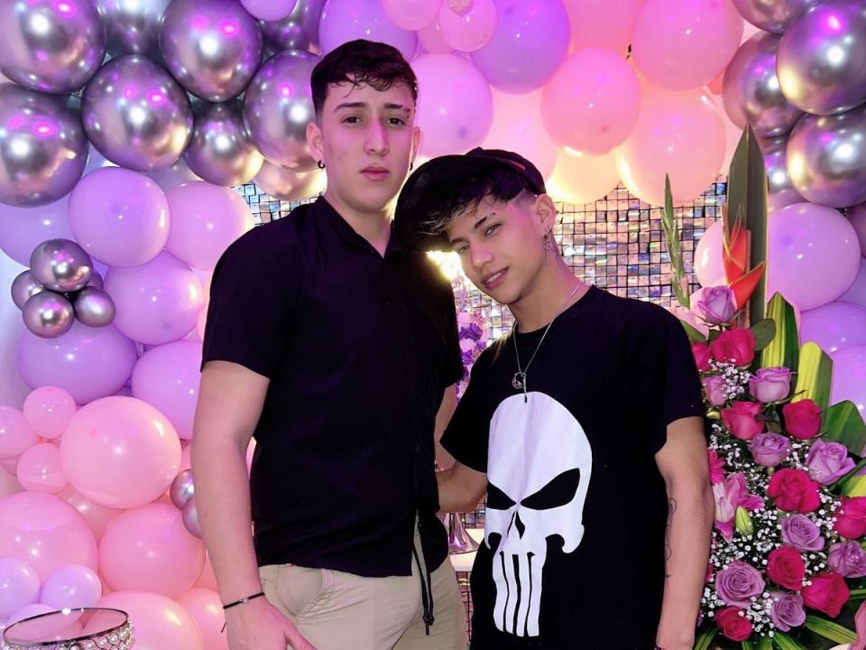Two TwinksHot with black shirts and short curly hair, standing in front of a pink and silver balloon wall. One has a skull shirt, the other looks surprised.