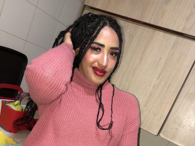 MichellDiaz with black braids, pink sweater, nose ring, red lipstick, smiling, hand in hair, kitchen, wooden cabinets, red basket with colorful items, sexy, cute, attractive.