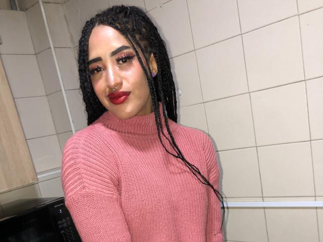 MichellDiaz with long black braids, pink knit sweater, red lipstick, nose ring, and bold eyeliner, smiling at the camera. Her tits are covered by the sweater.